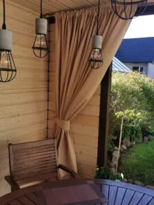 Didelė gelsvo atspalvio terasos užuolaida, plati užuolaida prie terasos sienos, surišta lauko užuolaida, metaliniai šviestuvai lauko terasoje.