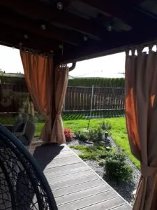Rusvos spalvos užuolaidos terasai, gražiai sutvarkyti terasa, apželdinta zona aplink terasą, akmenukai prie terasos.