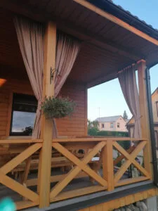 Gėlės pakabinamuose vazonuose, medinė terasa, lauko užuolaidos.