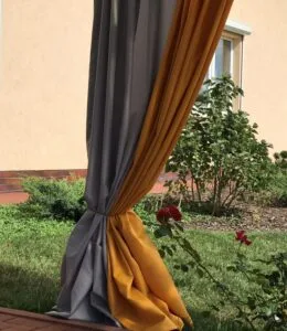 Geltona ir pilka užuolaida terasai, surišta ir pakabinta lauko terasoje, užuolaidos puošia terasą, pilkos ir kiaušinio trynio spalvos lauko užuolaida iš dviejų dalių.